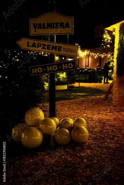 Obraz Valmiera,Latvia-November 30,2025-Festive outdoor Christmas scene with decorative signposts, glowing lights, evergreen trees and white ornaments on a warmly lit pathway.
