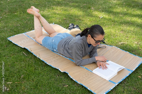 Fototapeta Woman relaxing and reading on grass mat in park.