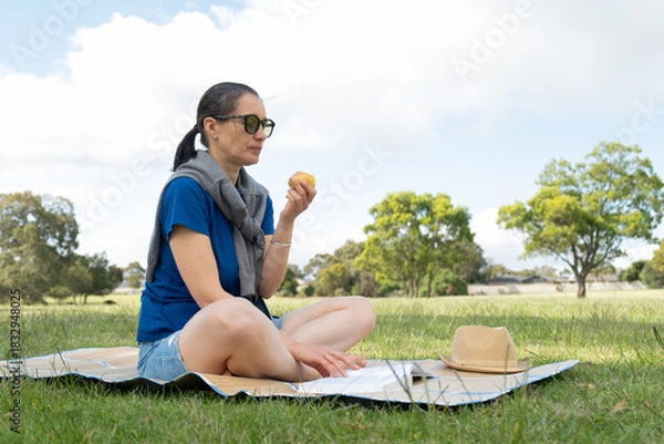 Fototapeta Woman enjoying picnic in sunny park, reading book and eating pear.