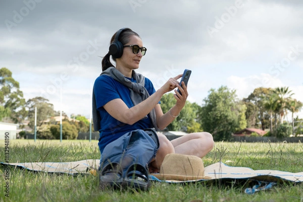 Fototapeta Woman relaxing in park wearing headphones and browsing on a smartphone.