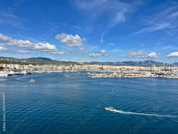 Obraz Scenic panoramic view of a coastal city with marina, yachts, and calm blue sea under a bright sky with scattered clouds, surrounded by hills and buildings