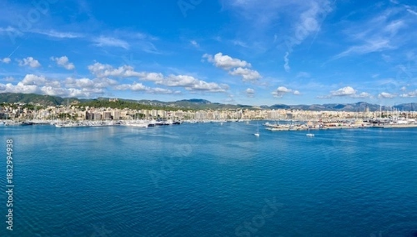 Obraz Scenic panoramic view of a coastal city with marina, yachts, and calm blue sea under a bright sky with scattered clouds, surrounded by hills and buildings