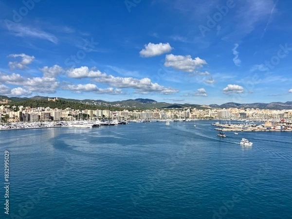Obraz Scenic panoramic view of a coastal city with marina, yachts, and calm blue sea under a bright sky with scattered clouds, surrounded by hills and buildings