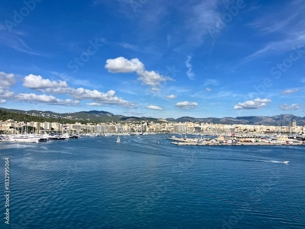 Obraz Scenic panoramic view of a coastal city with marina, yachts, and calm blue sea under a bright sky with scattered clouds, surrounded by hills and buildings