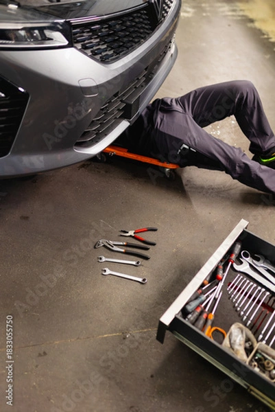 Fototapeta Mechanic lying underneath a raised vehicle in the workshop. Repair, inspection, and maintenance of the car underbody.