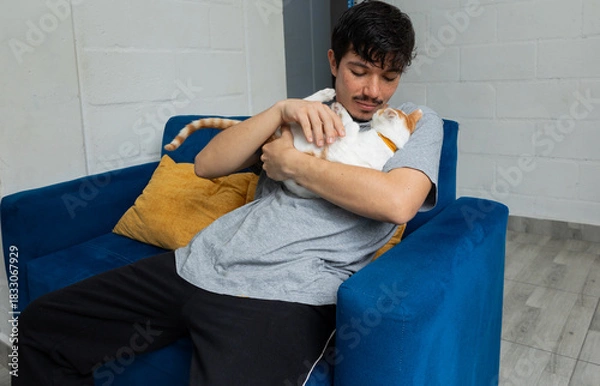 Obraz Man sitting on the sofa in his living room, petting and holding his cat, showing tenderness and connection with his pet at home