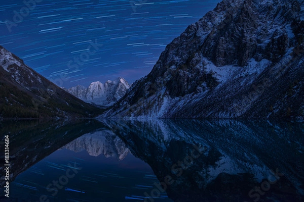 Obraz Stars motion are reflecting with bright trails on the smooth surface of clear mountain lake at the foot of sheer slopes covered with snow and high capped peaks with glaciers, Siberia, Altai, Shavla