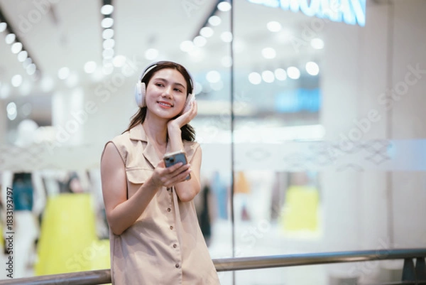Fototapeta A smiling woman enjoying music with headphones while holding her smartphone, surrounded by bright decorative lights in a lively atmosphere.