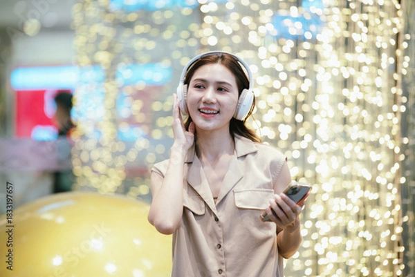 Fototapeta A smiling woman enjoying music with headphones while holding her smartphone, surrounded by bright decorative lights in a lively atmosphere.