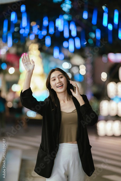 Fototapeta A smiling woman using her smartphone at night in a lively city setting, creating a warm and relaxed atmosphere with colorful blurred lights in the background.