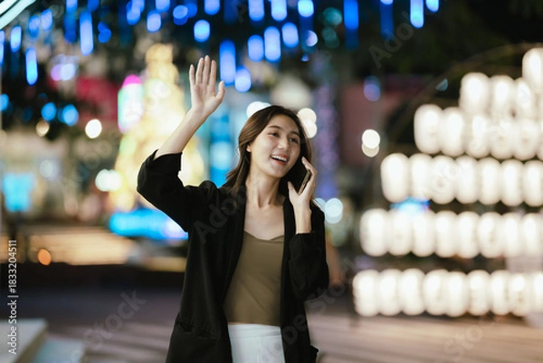 Fototapeta A smiling woman using her smartphone at night in a lively city setting, creating a warm and relaxed atmosphere with colorful blurred lights in the background.