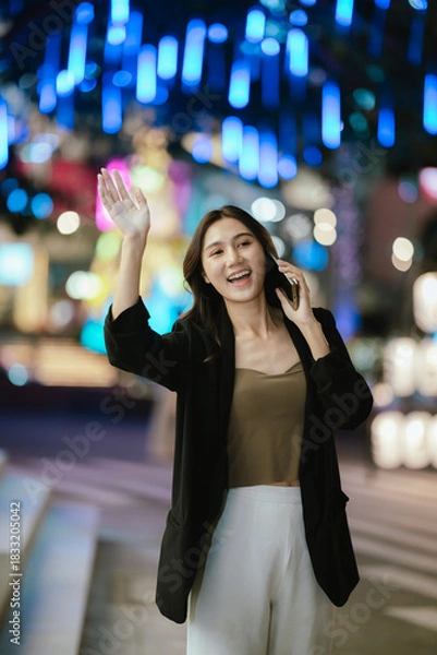 Fototapeta A smiling woman using her smartphone at night in a lively city setting, creating a warm and relaxed atmosphere with colorful blurred lights in the background.