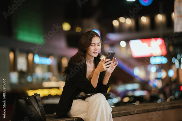 Fototapeta A smiling woman using her smartphone at night in a lively city setting, creating a warm and relaxed atmosphere with colorful blurred lights in the background.