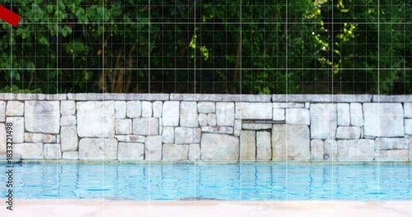 Fototapeta Showing pool water rippling in backyard by stone wall and green treeline, pale deck, red triangle