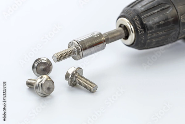 Obraz Close-up of a cordless drill with metal bit and three hex bolts resting on a white surface for hardware and DIY projects