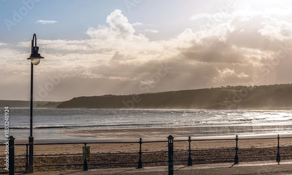 Fototapeta A promenade beside a beach with a headland in the distance and a cloudy sky.