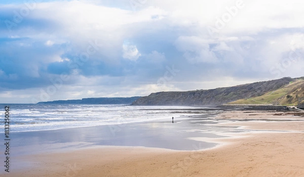 Fototapeta A sandy beach with a rough sea and headlands. A cloudy sky is above.