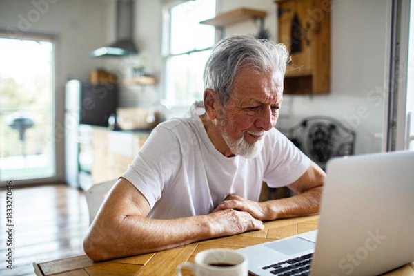 Fototapeta Senior man smiling while using laptop in home kitchen