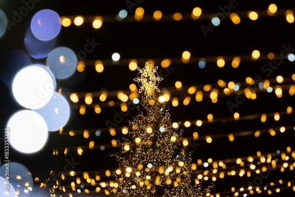 Fototapeta A festive outdoor scene featuring a Christmas tree adorned with golden lights, surrounded by strings of warm, glowing bokeh lights against a dark night sky
