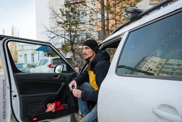 Fototapeta urban man exiting, individual emerges from car onto city sidewalk, man dressed casually with beanie departs vehicle to run brief city errands amid buildings and parked vehicles