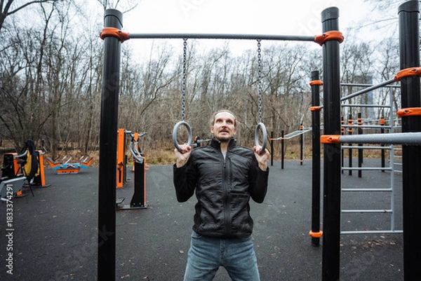 Fototapeta man practicing strength training in park, focused individual performing gymnastic ring exercises outdoors, man engaged in disciplined calisthenics using rings in urban park setting