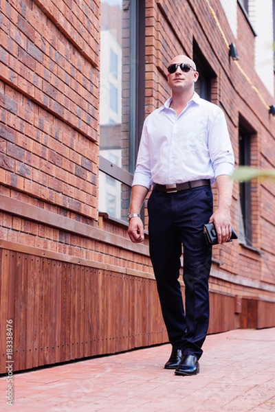 Obraz bald strong businessman full height going to work next duilding from red brick . shirt dark blue trousers, boots. looking straight. spring, summer