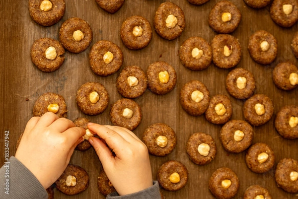 Fototapeta Top view of child's hands pressing hazelnuts into raw cookie dough. Preparing traditional Christmas treats