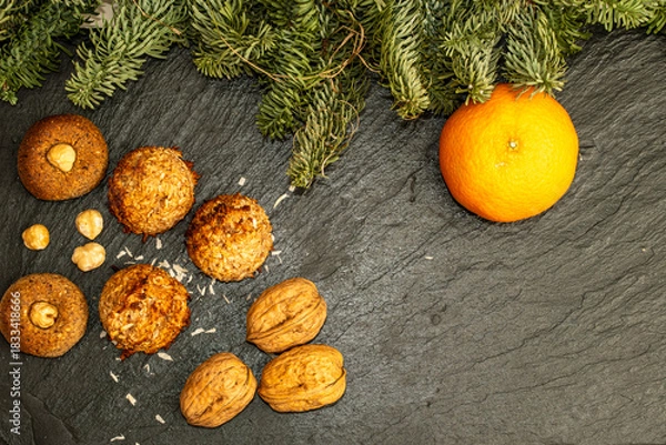 Fototapeta Overhead shot of Christmas baking treats including coconut cookies and walnuts on a black slate background decorated with fir branches and a fresh orange