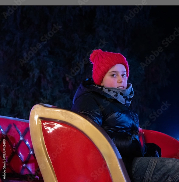 Obraz A portrait of a Boy in a red hat on a Christmas Carousel in an Evening Park