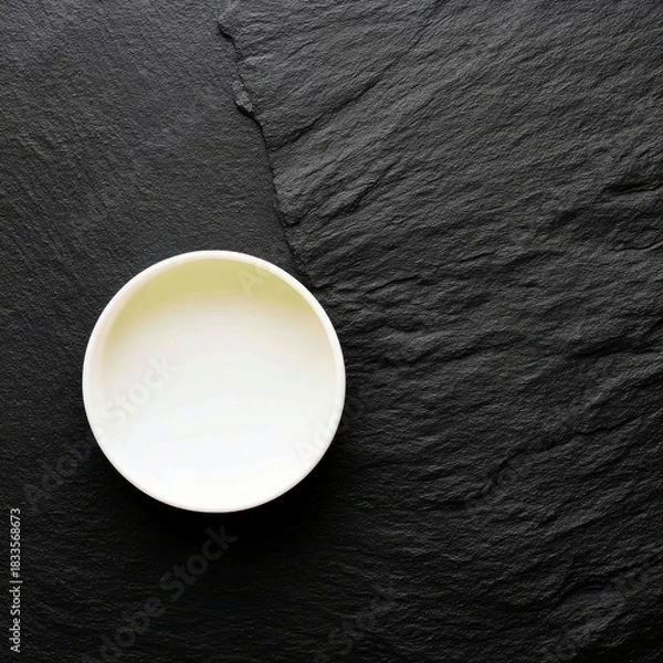 Fototapeta An overhead shot of an empty white bowl on a textured black slate stone surface. The bowl is centered, and the lighting is soft.