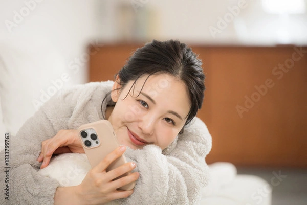 Obraz 冬のモコモコした部屋着で寝転がるスマホを見る若い女性カメラ目線