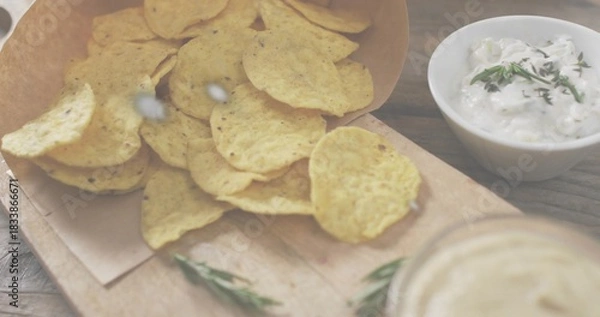 Fototapeta Spilling corn tortilla chips from paper cone onto wooden board on rustic tabletop, with herb dip