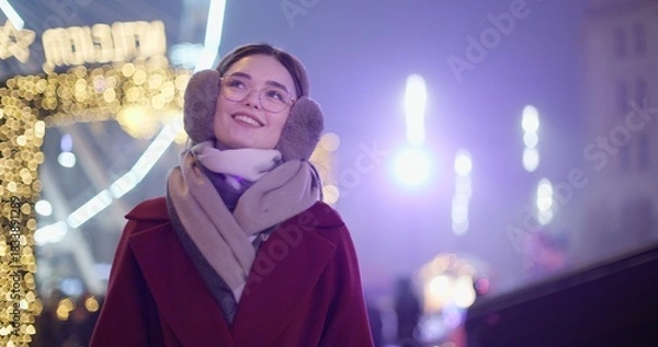 Obraz A young beautiful girl in a red coat walks in the evening at the New Year's Christmas fair against the backdrop of a Ferris wheel among lights and garlands and admires the decorations