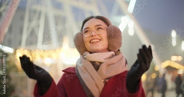 Obraz A young beautiful girl in a red coat walks in the evening at the New Year's Christmas fair against the backdrop of a Ferris wheel among lights and garlands and admires the decorations