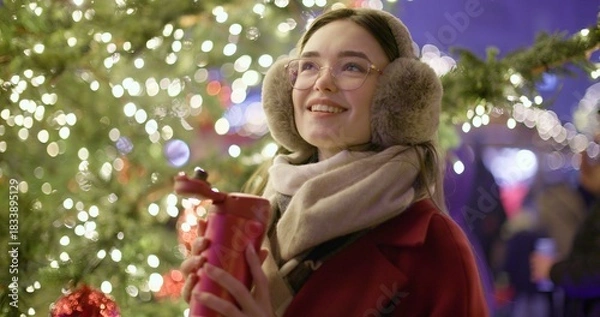 Obraz A young beautiful girl in a red coat walks in the evening at the European Christmas Market and holds a thermos cup, standing against the backdrop of a Christmas tree with garlands and illuminations.