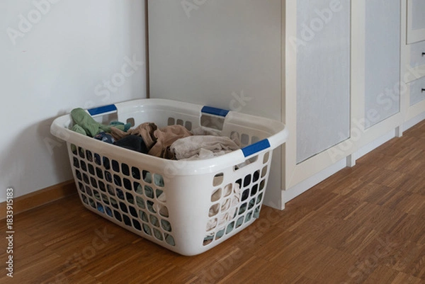 Fototapeta A cluttered laundry basket filled with assorted clothing, positioned neatly against a wall in a modern interior with wooden flooring and a beige wardrobe