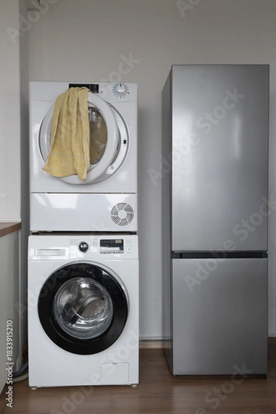 Fototapeta Stacked washing machine and dryer beside a tall grey refrigerator in a laundry room with a yellow towel hanging on the dryer