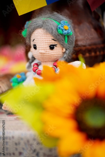 Obraz Close-up of a handmade crocheted doll with grey braids and flower accessories, partially obscured by a bright orange sunflower in the foreground