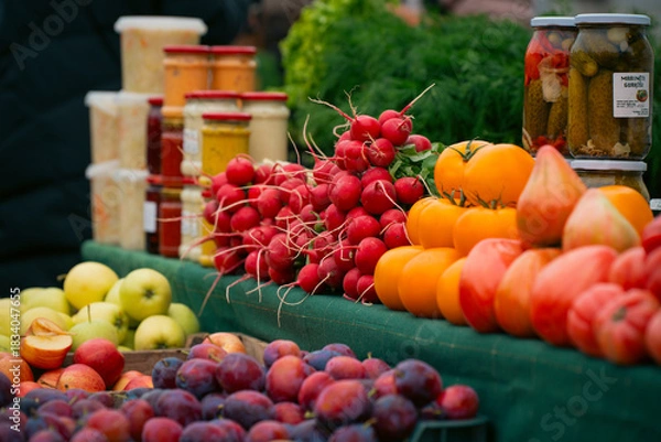 Obraz Fresh vegetables and fruits displayed at a local market, including radishes, tomatoes, apples, and plums, with jars of homemade preserves in the background