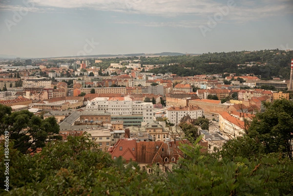 Fototapeta Panoramic city view of Brno from an observation point near Špilberk Castle, showing red rooftops, urban districts and green hills under a soft summer sky in the Czech Republic.