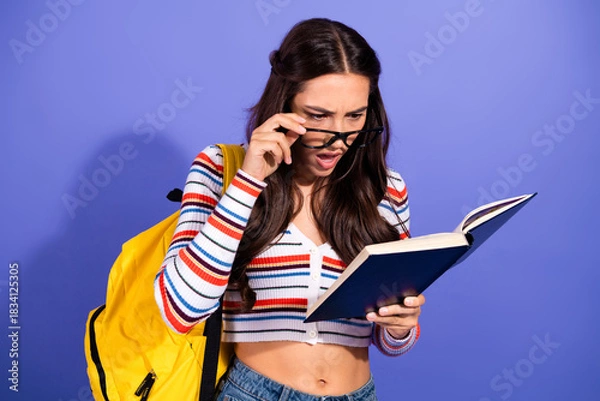 Fototapeta Young female student reads a book with glasses and a yellow backpack against a purple background