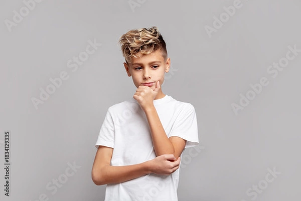 Obraz Thoughtful young boy with trendy blond hair touching chin while looking down, wearing white T-shirt and posing in studio, isolated on gray background in modern portrait