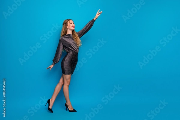 Obraz Stylish woman in a shiny dress poses confidently against a blue studio background for stock photography with glamour and fashion appeal