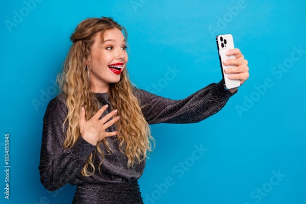 Obraz Joyful glamorous woman takes a selfie with a smartphone in a glittering dress against a bright blue background