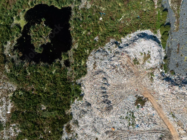 Fototapeta Aerial view showing a stark contrast between a natural wetland with a dark pond and a nearby landfill area.