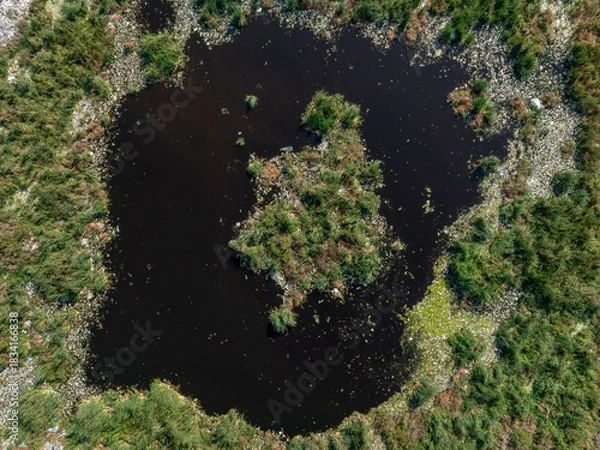 Obraz Aerial view showing a stark contrast between a natural wetland with a dark pond and a nearby landfill area.