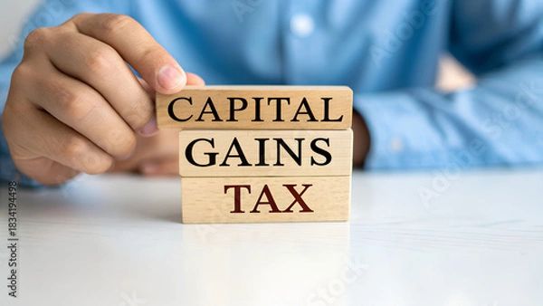 Fototapeta A man is holding wooden blocks with the words capital gains tax written on them on a white table