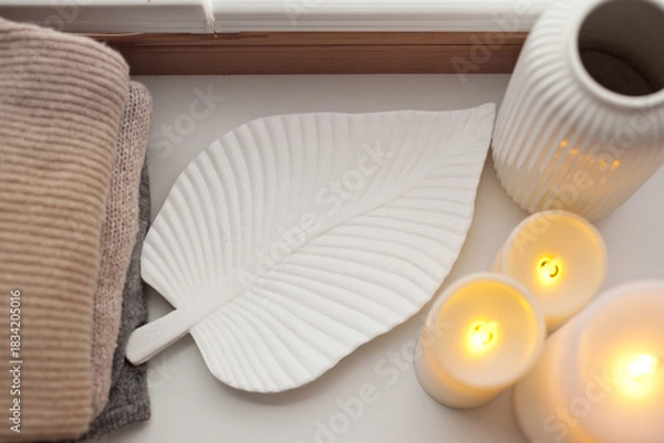 Obraz Pile of beige knitted sweaters, wooden tray, candles and vase over white background top view