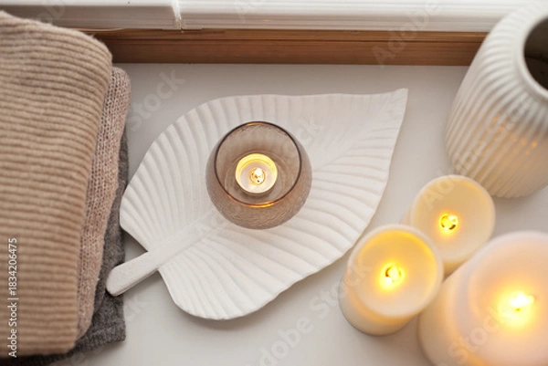 Obraz Pile of beige knitted sweaters, wooden tray, candles and vase over white background top view