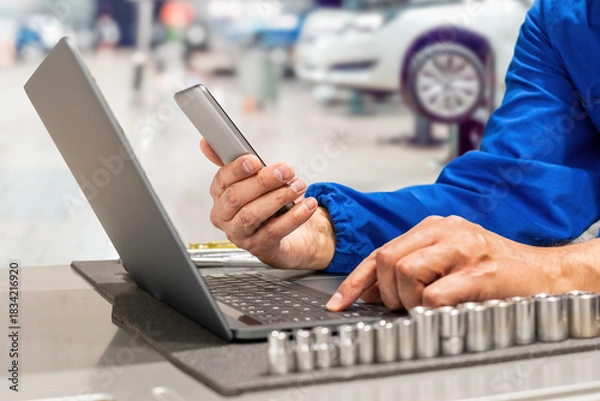 Fototapeta Close-up of hands of manager at the auto repair shop  in a blue uniform a car engine, engaging with his mobile phone in a garage environment, embodying satisfaction and modern technology at work